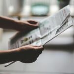 Hands holding a newspaper in a soft-lit, indoor environment, focusing on a relaxed reading moment.